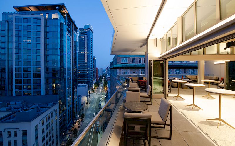A modern cityscape at dusk with tall buildings and a lit balcony area furnished with tables and chairs overlooking the streets below.
