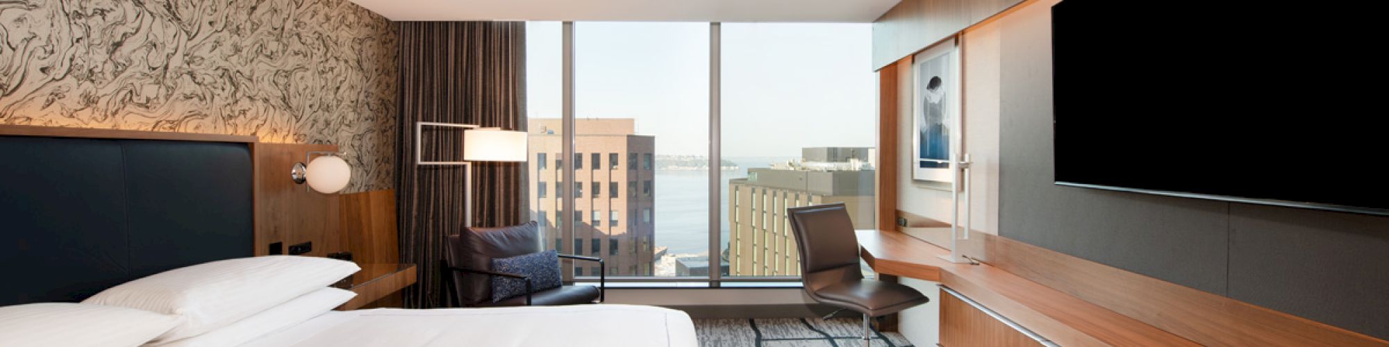 A modern hotel room with a large bed, a desk, a chair, a flat-screen TV, and a window offering a view of nearby buildings and the water.